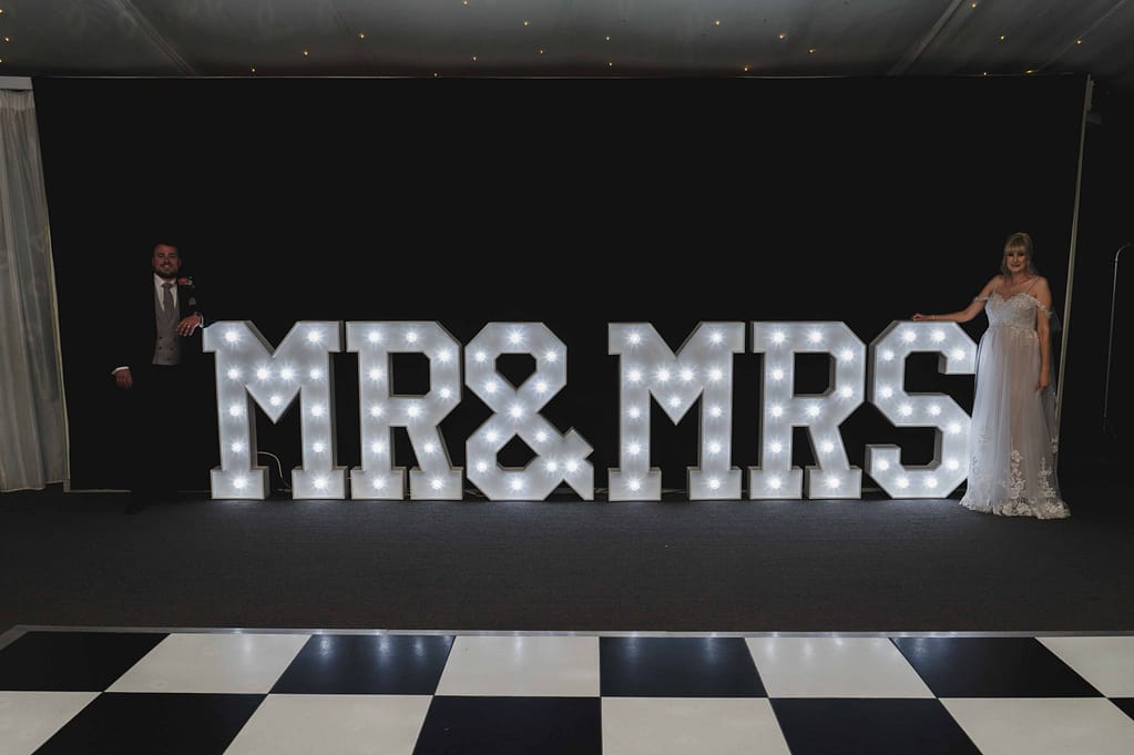 Bride & Groom standing either end of a neon Mr&MRs sign in the marque at Braxted Park wedding venue