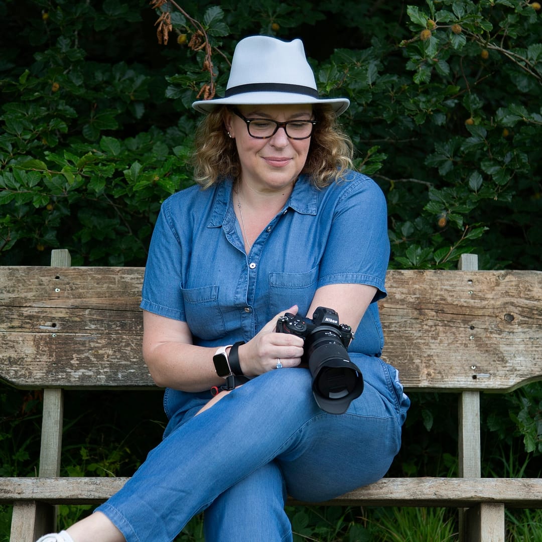 Catherine Brooks Photography - Catherine a lady in a denim jumpsuit, sat on a wooden bench with her camera. She is wearing a grey fedora hat and black glasses