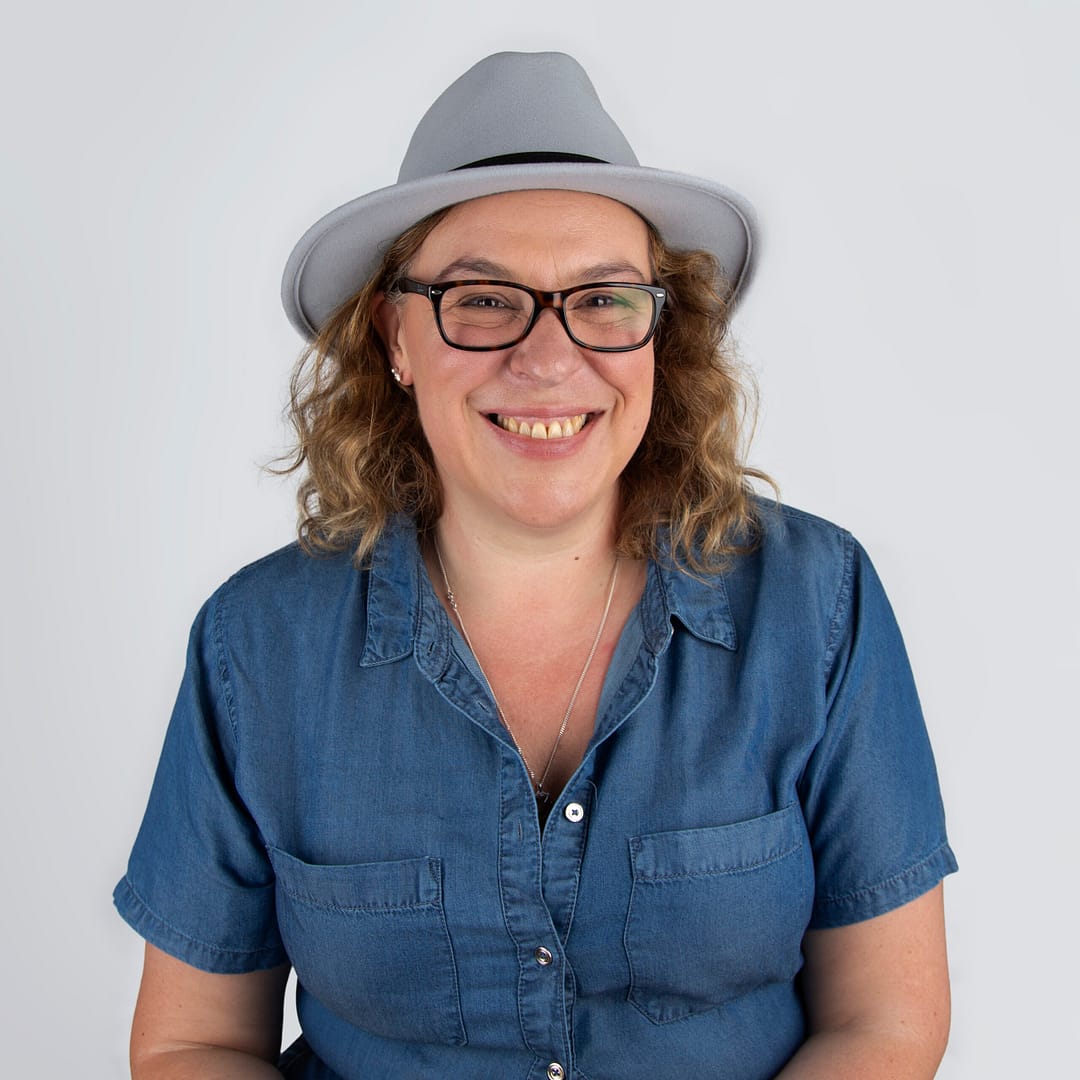 Catherine Brooks Photography - Image of Catherine a lady wearing a denim shirt with curly mouse brown hair to the shoulder and glasses. She has a grey fedora hat on her hat and black glasses and is smiling