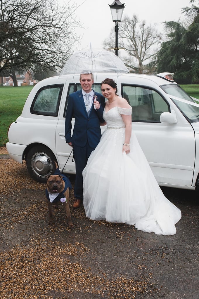 Bride & Groom under a brolly standing in front of a white taxi with dog